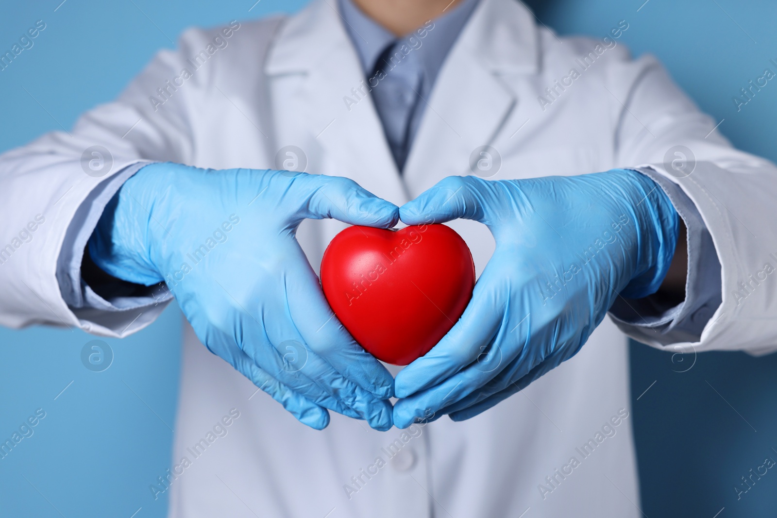 Photo of Doctor wearing medical gloves holding decorative heart on light blue background, closeup