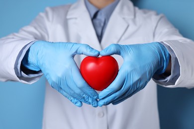 Doctor wearing medical gloves holding decorative heart on light blue background, closeup