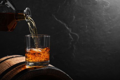 Photo of Pouring whiskey into glass from bottle on wooden barrel against grey background, space for text