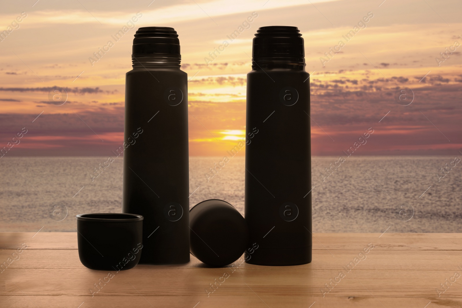 Image of Thermoses on wooden table near sea under sunset sky