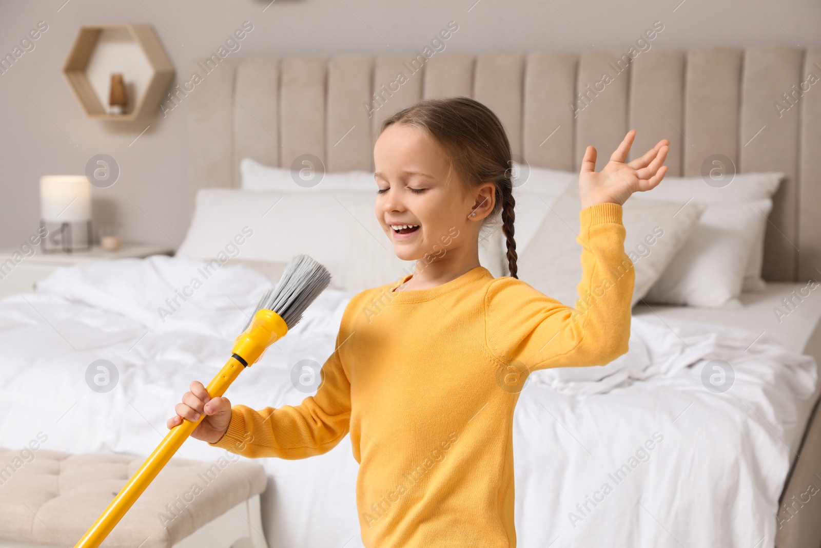 Photo of Cute little girl with broom singing while cleaning in bedroom