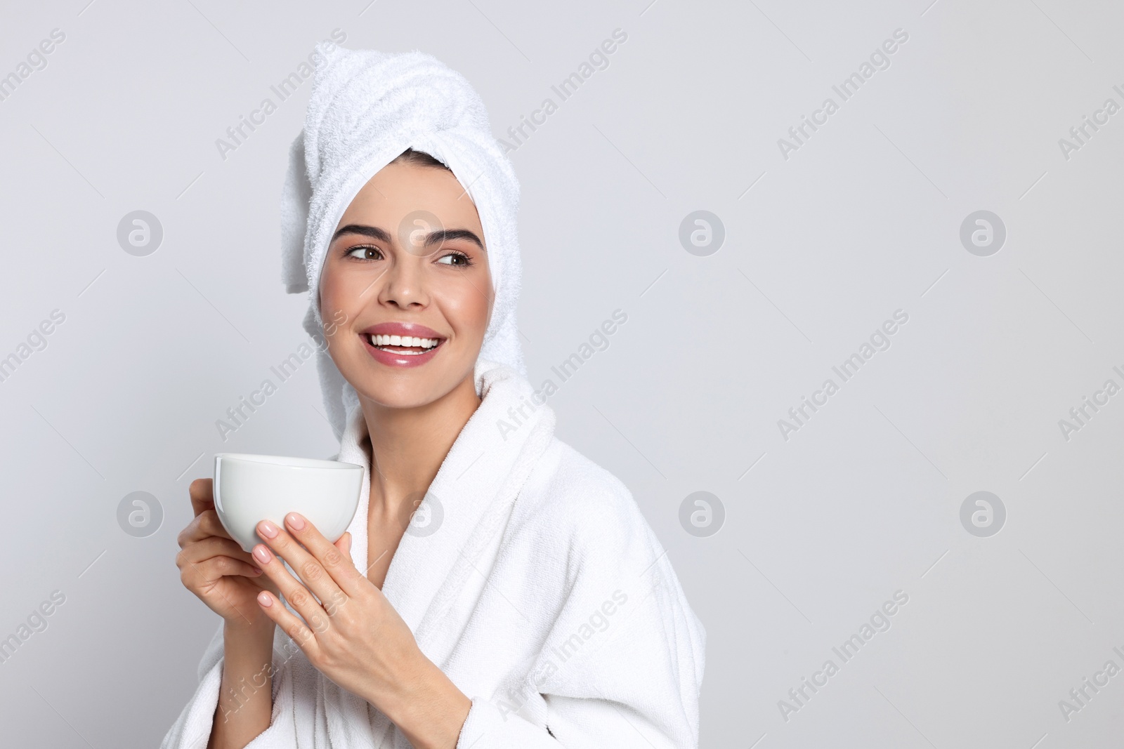 Photo of Young woman in bathrobe with towel and cup of drink on light grey background, space for text. Spa treatment