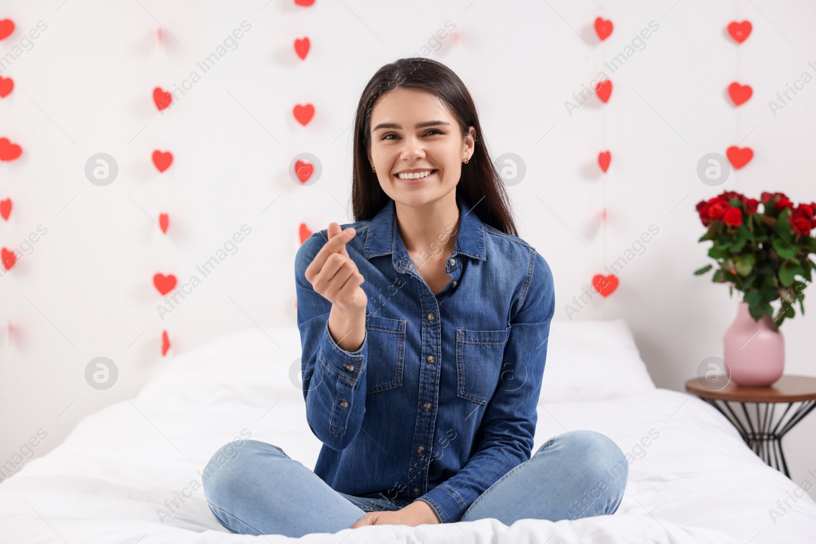 Photo of Beautiful young woman showing heart gesture in bedroom, view from camera. Valentine's day celebration in long distance relationship