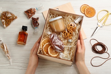 Woman holding stylish gift set at white wooden table, top view