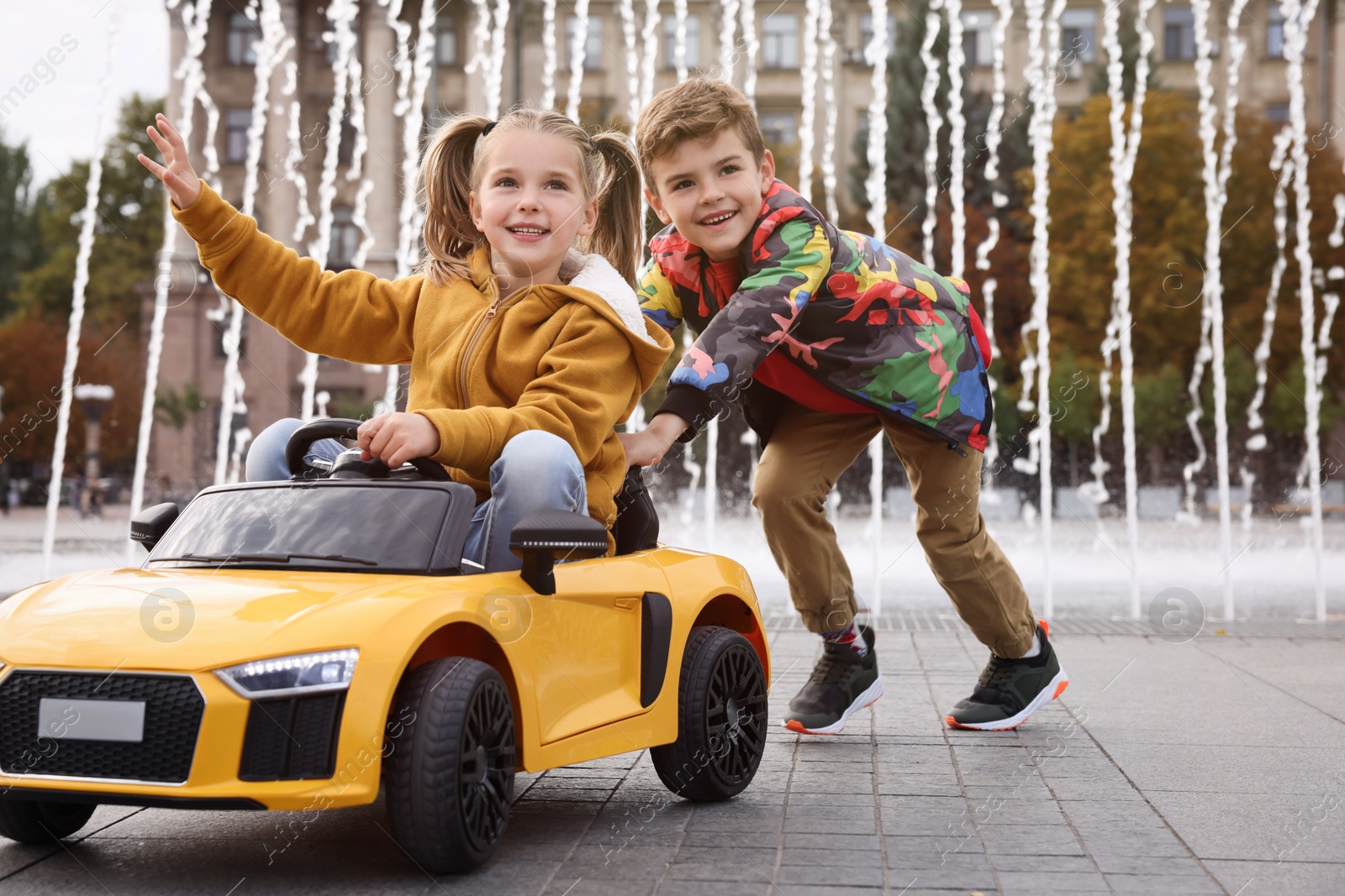 Photo of Cute boy pushing children's car with little girl near fountain on city street