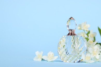 Photo of Aromatic perfume in bottle and beautiful jasmine flowers on light blue background, closeup. Space for text