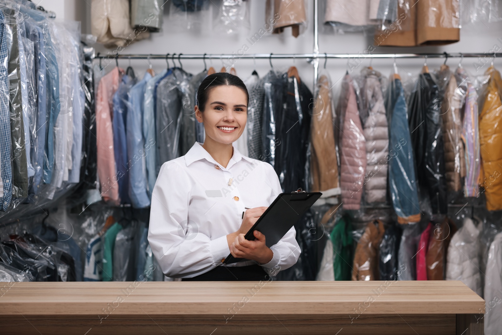 Photo of Dry-cleaning service. Happy worker with clipboard at counter indoors