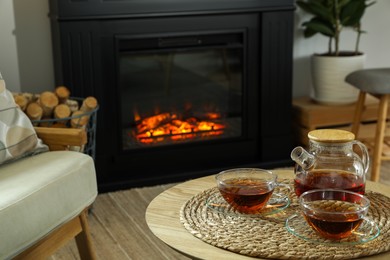 Teapot and cups of drink on coffee table near stylish fireplace in cosy living room. Interior design