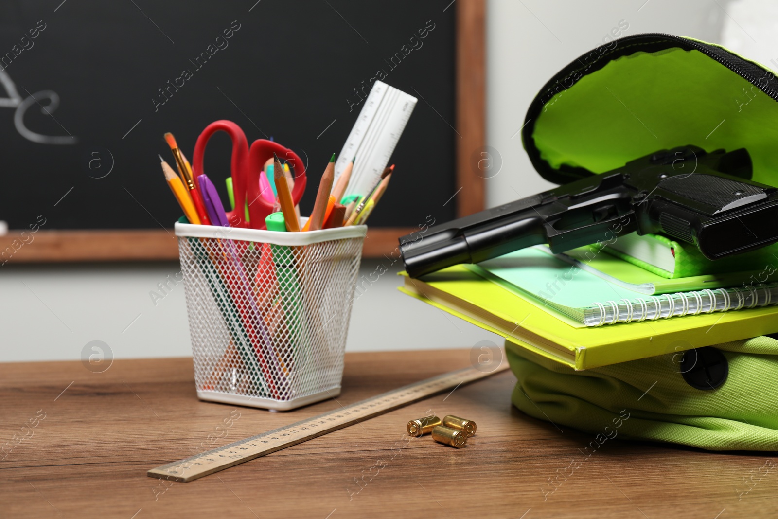 Photo of Gun, bullets and school stationery on wooden table near blackboard indoors