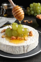 Photo of Pouring honey onto brie cheese served with grape and cashew nuts on dark grey table, closeup