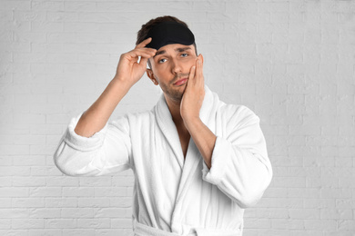 Sleepy young man in bathrobe and eye sleeping mask near white brick wall