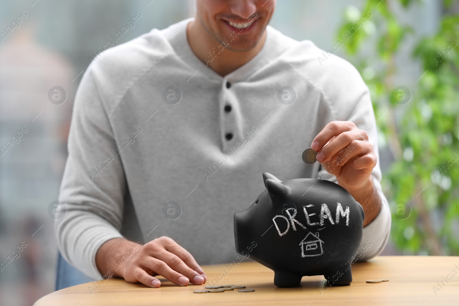 Photo of Man putting money into piggy bank with word DREAM at table, closeup