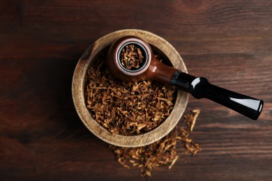 Smoking pipe and bowl of dry tobacco on wooden table, top view