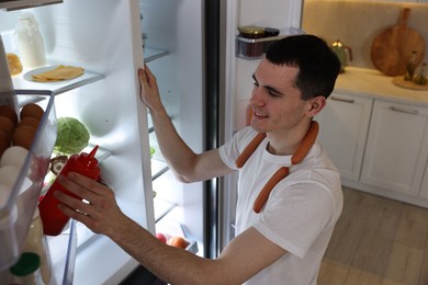 Photo of Happy man with sausages taking ketchup out of refrigerator in kitchen
