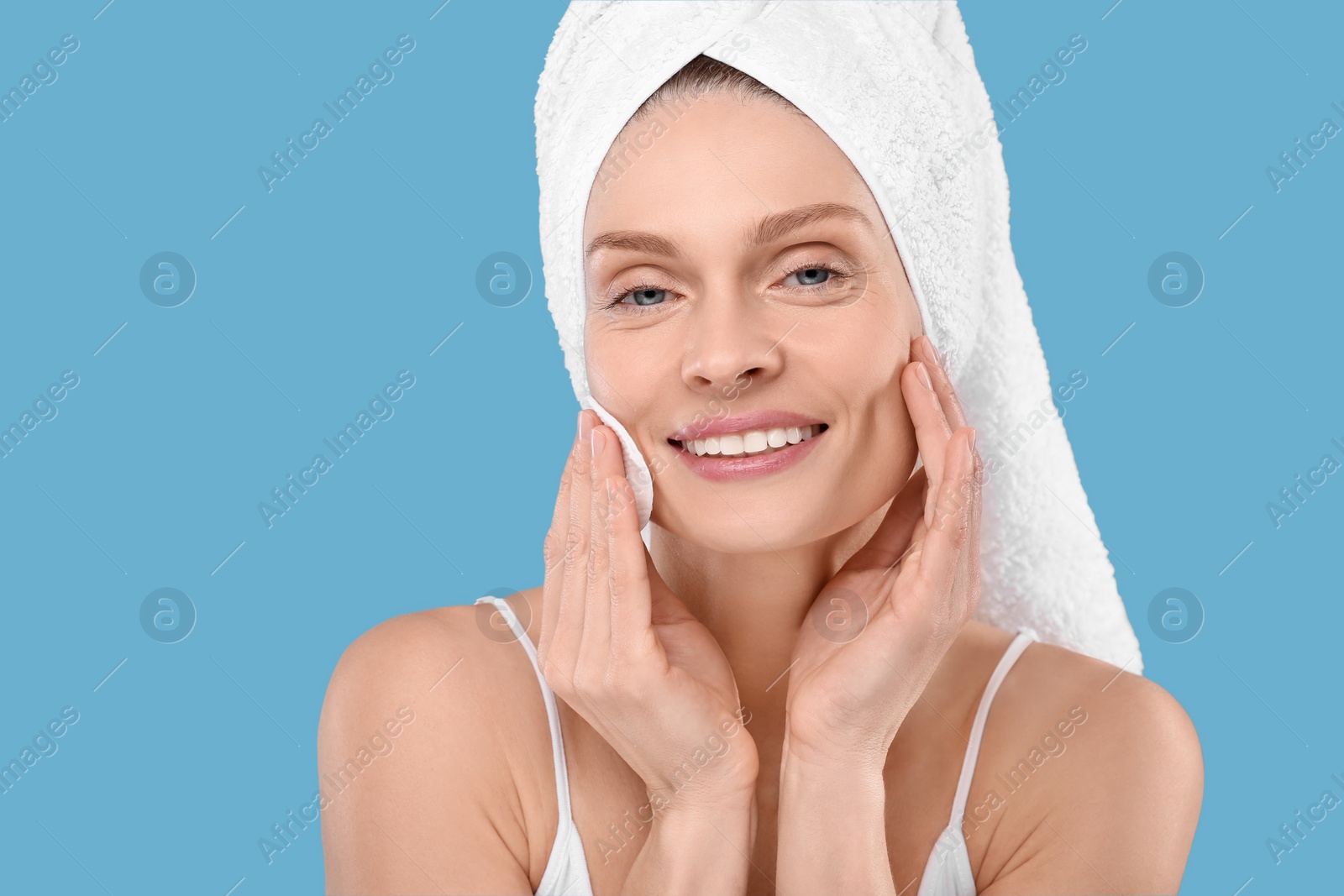 Photo of Beautiful woman in terry towel removing makeup with cotton pad on light blue background