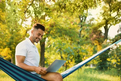 Young man with laptop in comfortable hammock at green garden