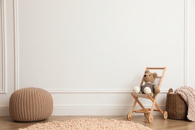 Toy stroller with bear, pouf and wicker basket near white wall in child room. Interior design