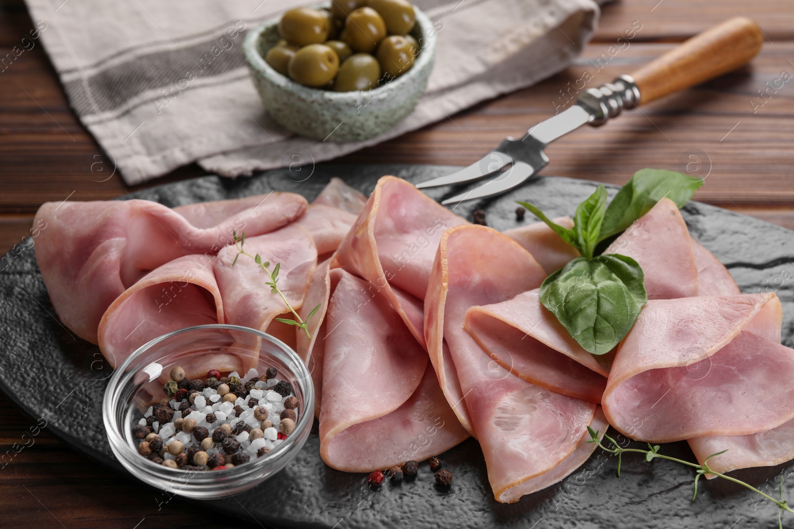 Photo of Tasty ham with basil, sea salt, peppercorns and carving fork on wooden table, closeup