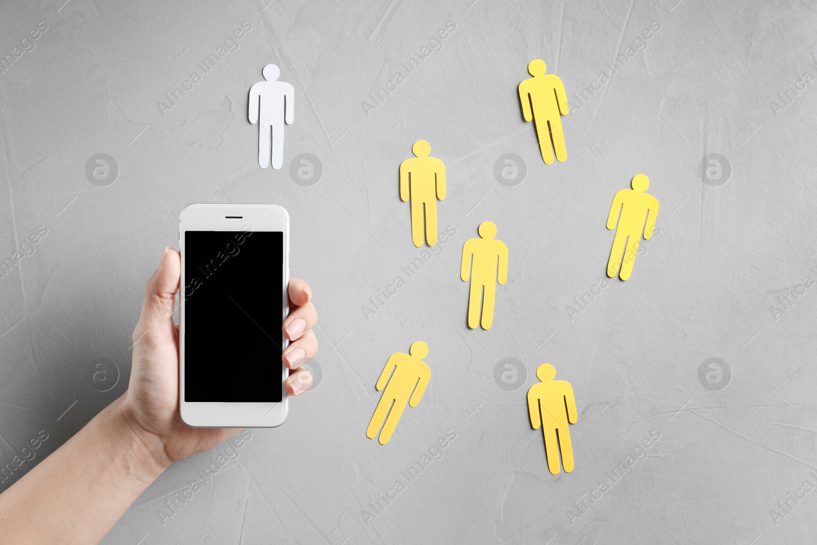 Photo of Woman holding smartphone over grey table with paper people, top view. Loneliness concept