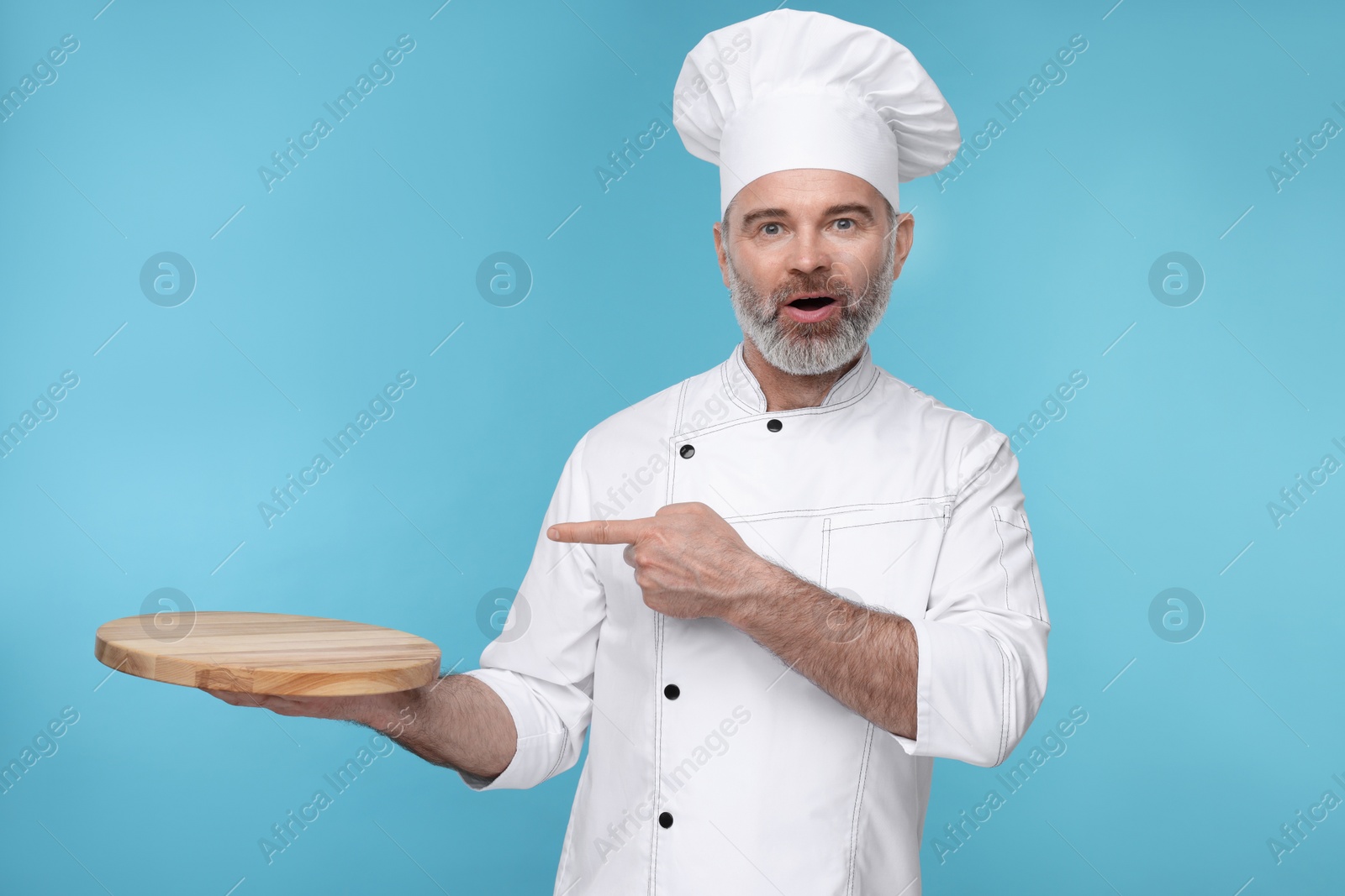 Photo of Surprised chef in uniform pointing at wooden board on light blue background