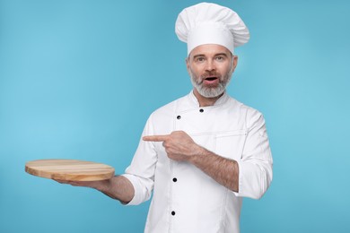 Surprised chef in uniform pointing at wooden board on light blue background