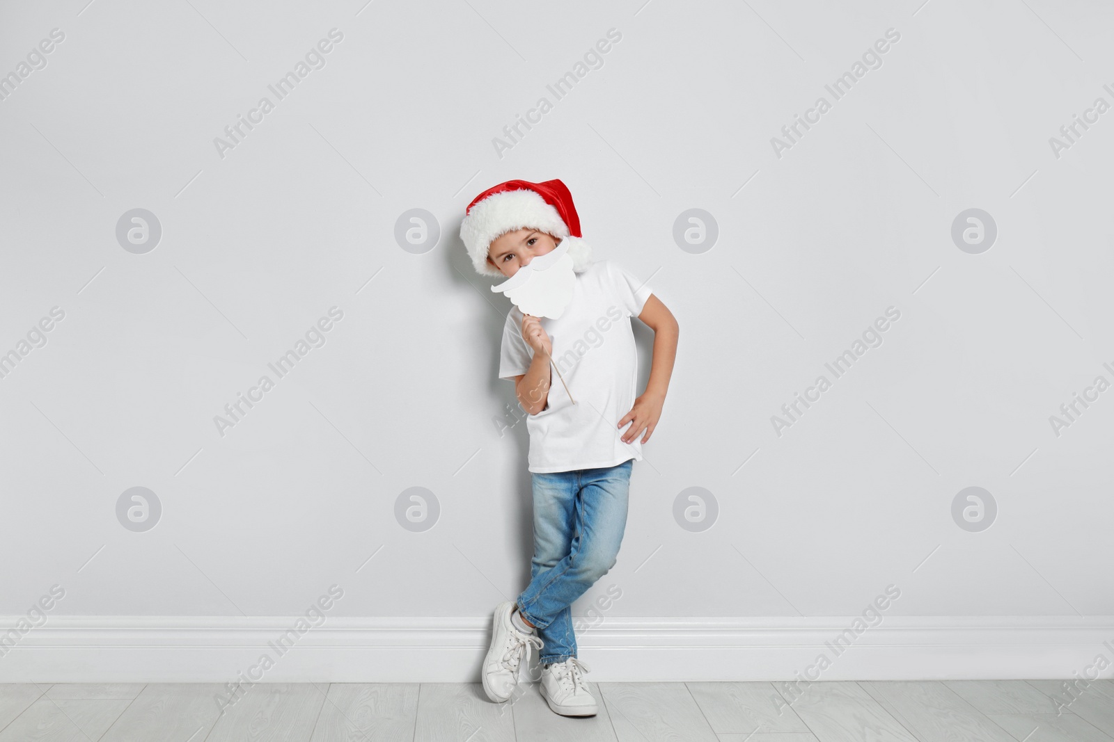 Image of Cute little boy with Santa hat and white beard prop near white wall. Christmas celebration