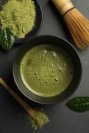 Photo of Cup of fresh matcha tea, bamboo whisk, spoon and green powder on black table, flat lay