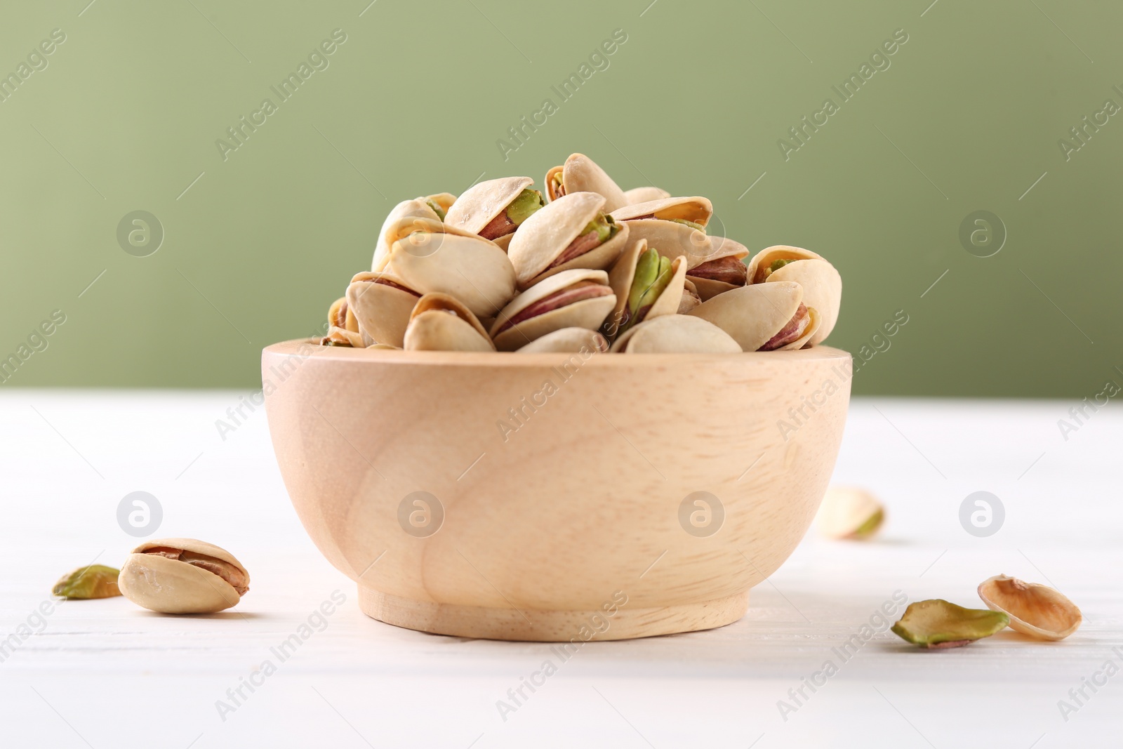 Photo of Tasty pistachios in bowl on white wooden table against olive background, closeup