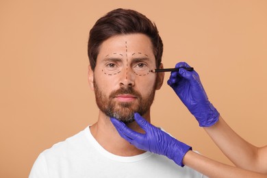 Doctor drawing marks on man's face for cosmetic surgery operation against beige background