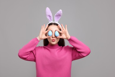 Photo of Happy woman in bunny ears headband holding painted Easter eggs near her eyes on grey background