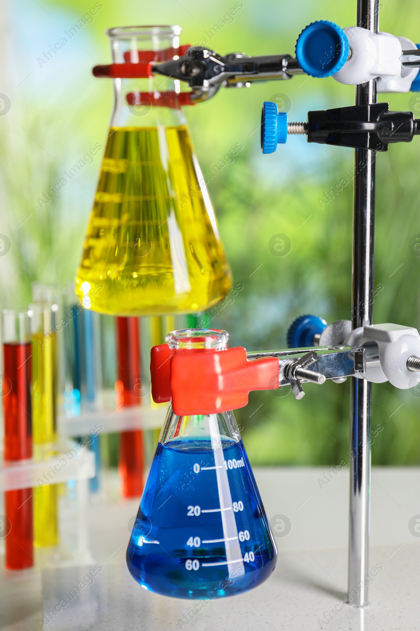 Photo of Retort stand and laboratory flasks with liquids on blurred background, closeup