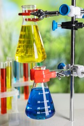 Photo of Retort stand and laboratory flasks with liquids on blurred background, closeup