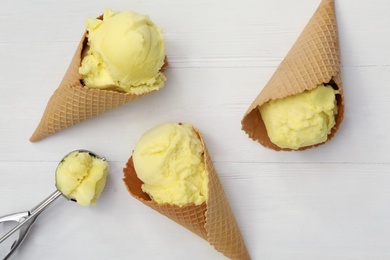 Photo of Yellow ice cream in wafer cones and scoop on white wooden table, flat lay