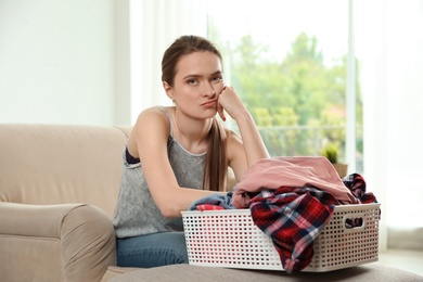 Photo of Upset young woman with basket of clean laundry on sofa in room