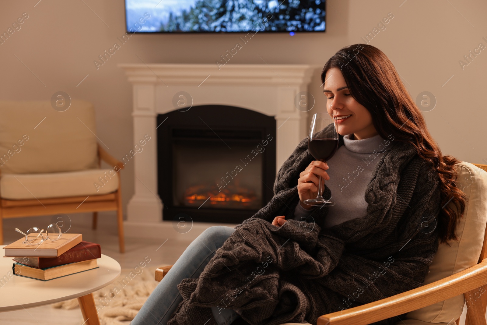 Photo of Young woman with glass of wine resting in armchair near fireplace at home