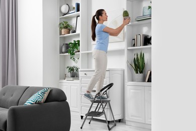Woman on ladder with houseplant near shelves at home