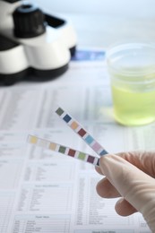 Photo of Doctor holding urine test strips near documents at table, closeup