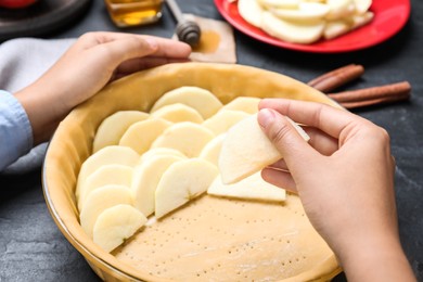 Photo of Woman putting apple slices into dish with raw dough at black table, closeup. Baking pie