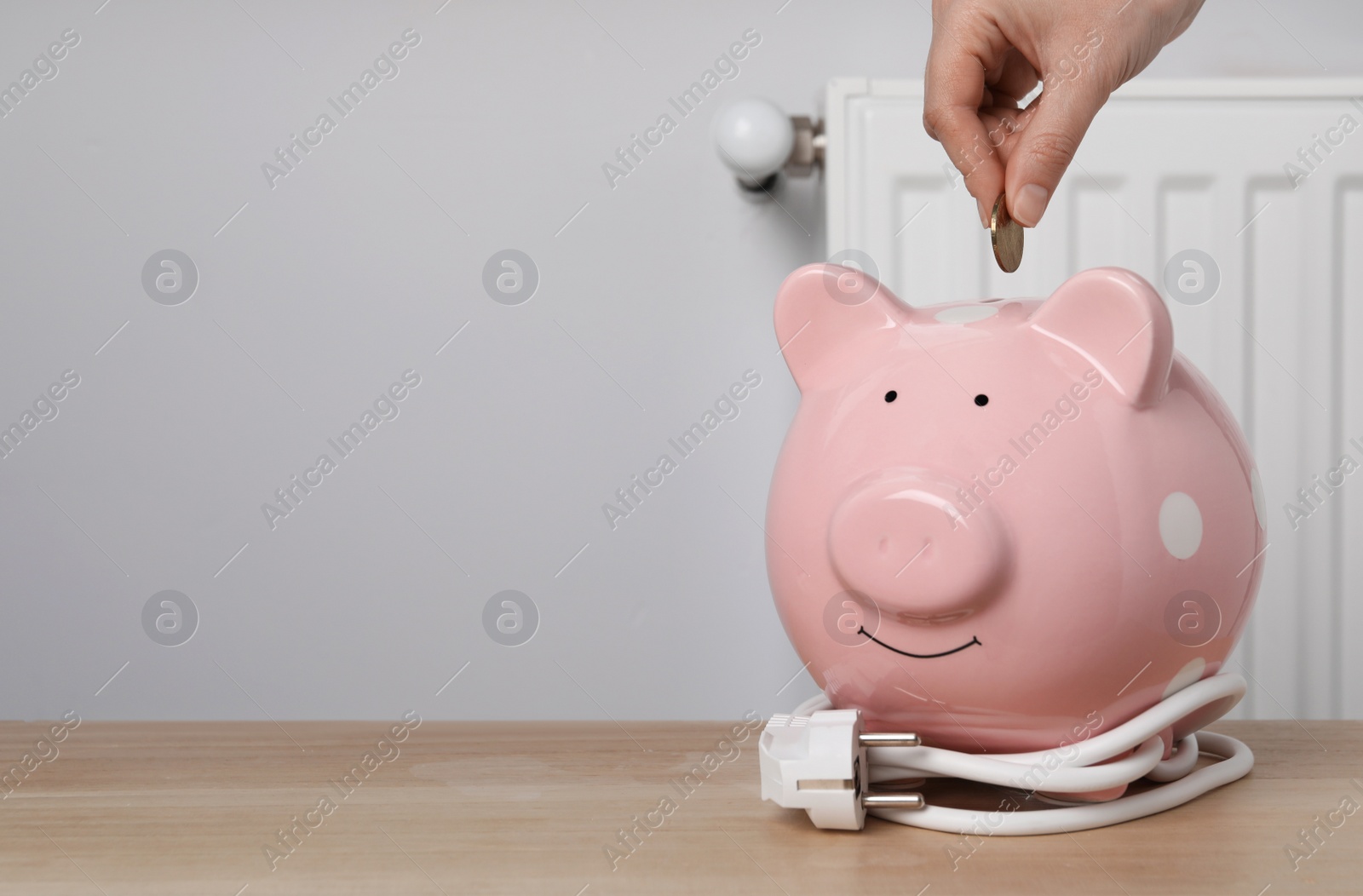 Photo of Woman putting coin into piggy bank at wooden table near heating radiator, closeup. Space for text