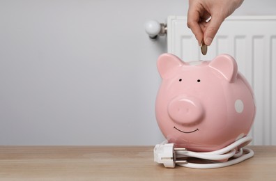 Woman putting coin into piggy bank at wooden table near heating radiator, closeup. Space for text