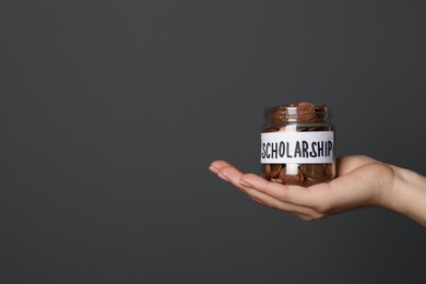 Photo of Woman holding glass jar of coins against dark grey background, closeup with space for text. Scholarship concept