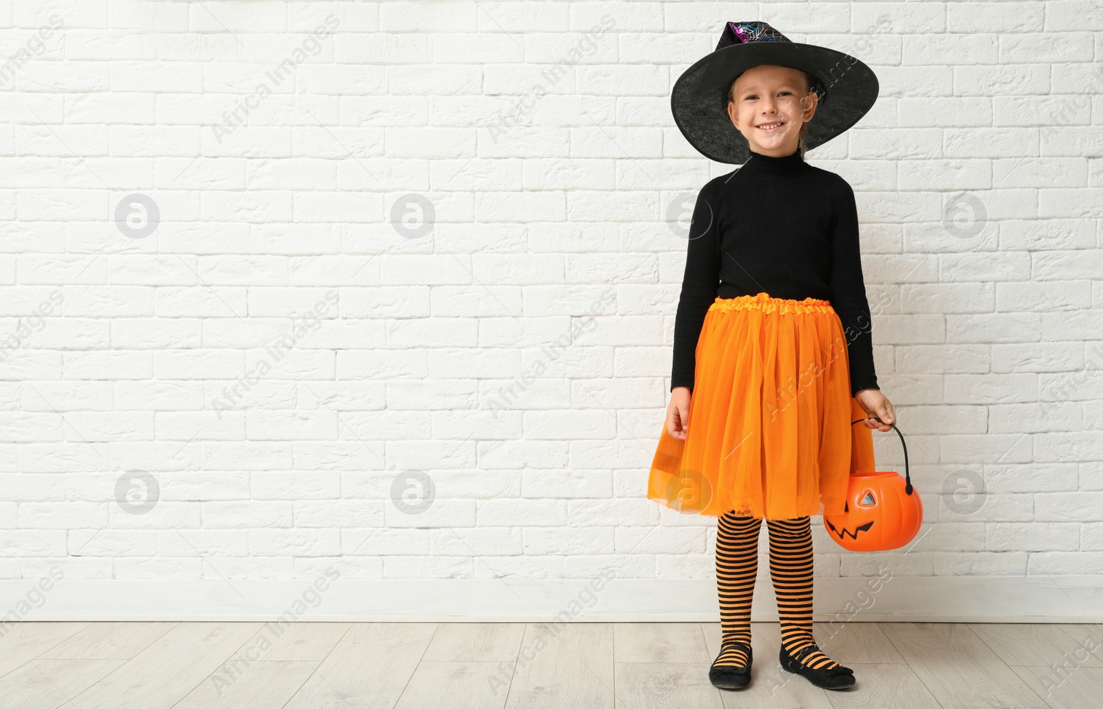 Photo of Cute little girl with pumpkin candy bucket wearing Halloween costume near white brick wall. Space for text