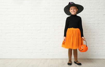 Photo of Cute little girl with pumpkin candy bucket wearing Halloween costume near white brick wall. Space for text