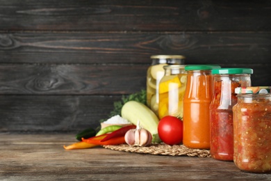 Photo of Jars with pickled products and fresh vegetables on wooden table against black background. Space for text
