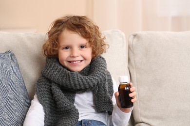 Photo of Cute boy holding bottle with cough syrup on sofa indoors. Effective medicine