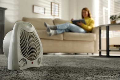 Photo of Woman reading book in living room, focus on electric fan heater