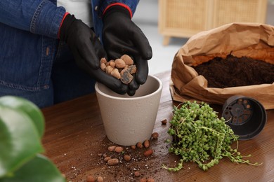 Photo of Woman in gloves filling flowerpot with drainage at wooden table indoors, closeup. Transplanting houseplants