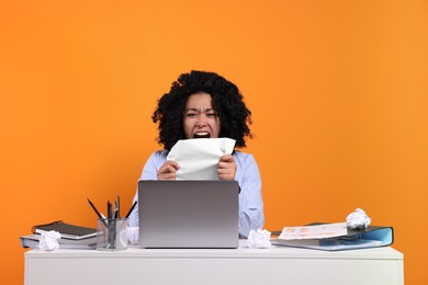 Stressful deadline. Emotional woman holding paper and shouting at white desk against orange background
