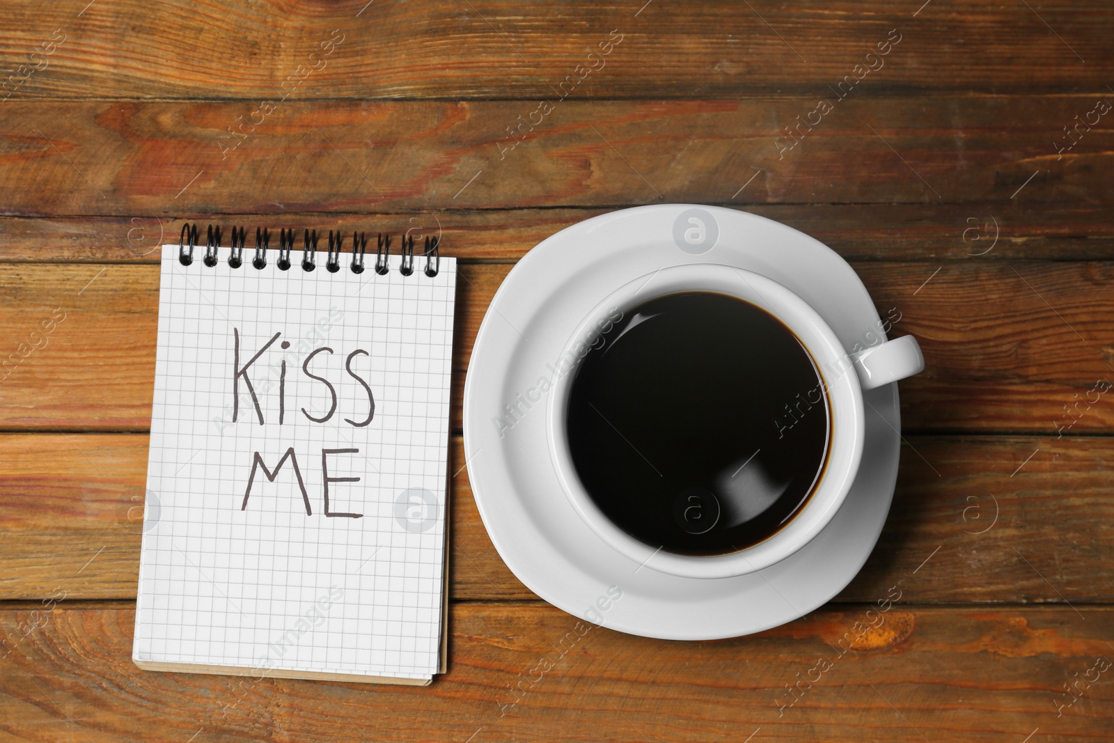 Photo of Notebook with phrase Kiss Me and cup of coffee on wooden table, flat lay