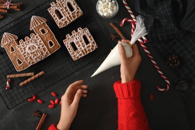 Photo of Woman decorating gingerbread house parts with icing at black table, top view